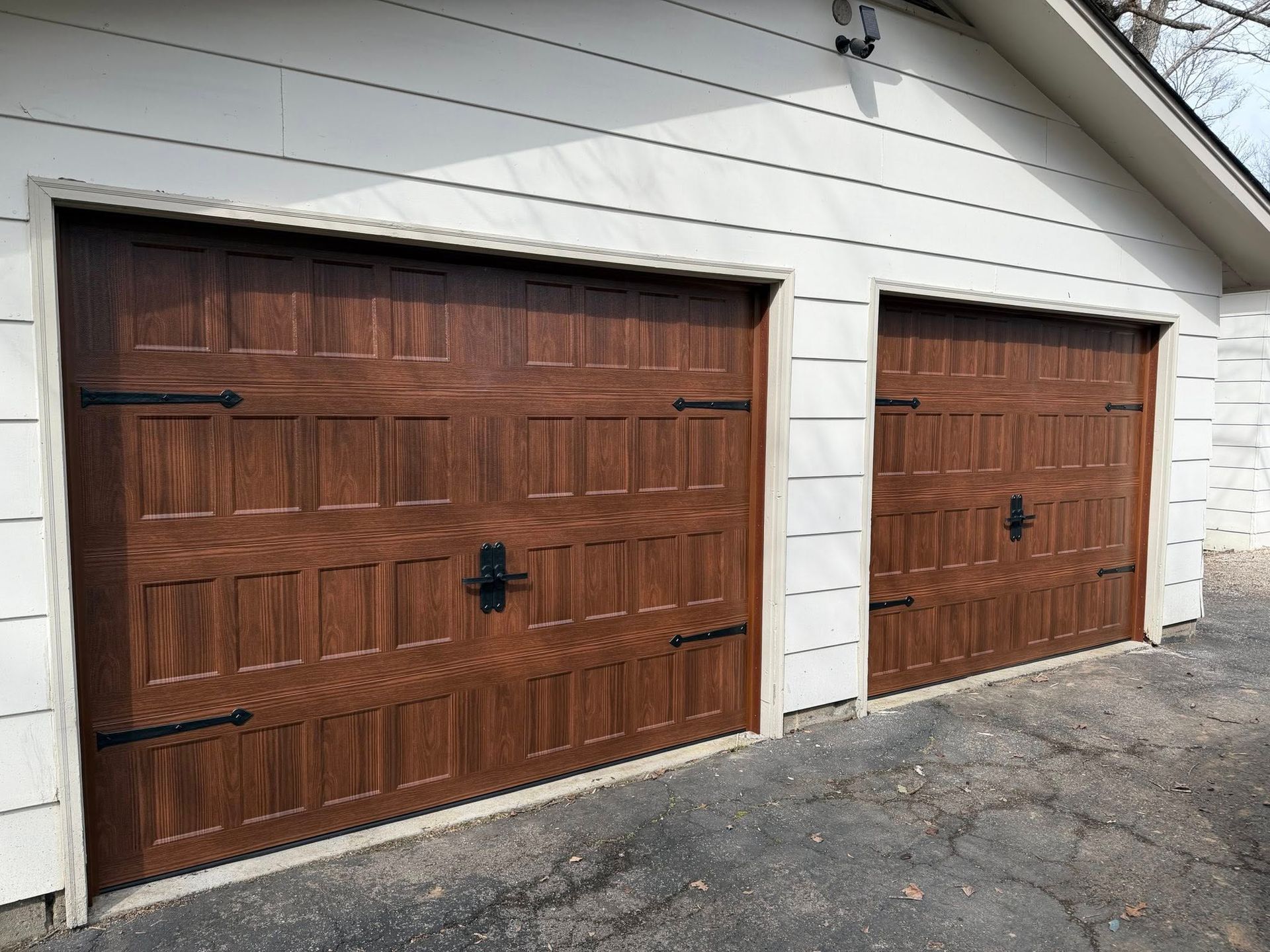 Two brown garage doors with black hardware, on a white house, set on a driveway.