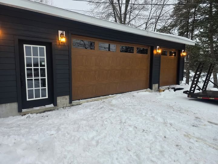 Brown garage doors and a white-framed door on a dark gray building, snow on the ground.