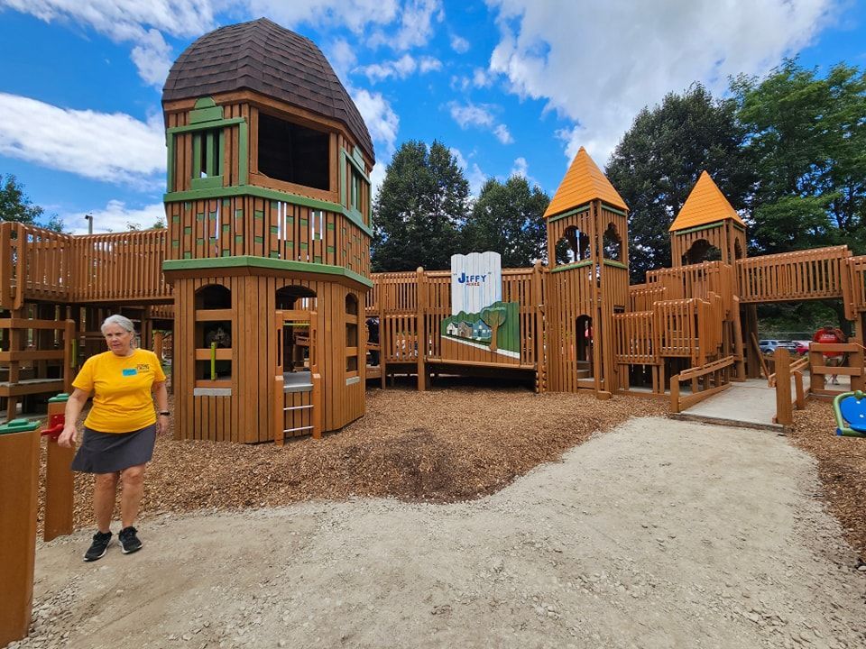 A woman in a yellow shirt is standing in front of a wooden playground.