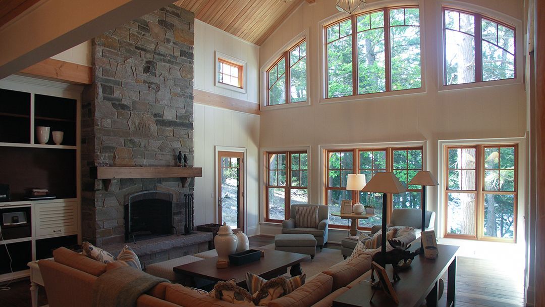 Large living room at a cottage, shows wide  curved windows  overlooking the lake, and a soaring fireplace,