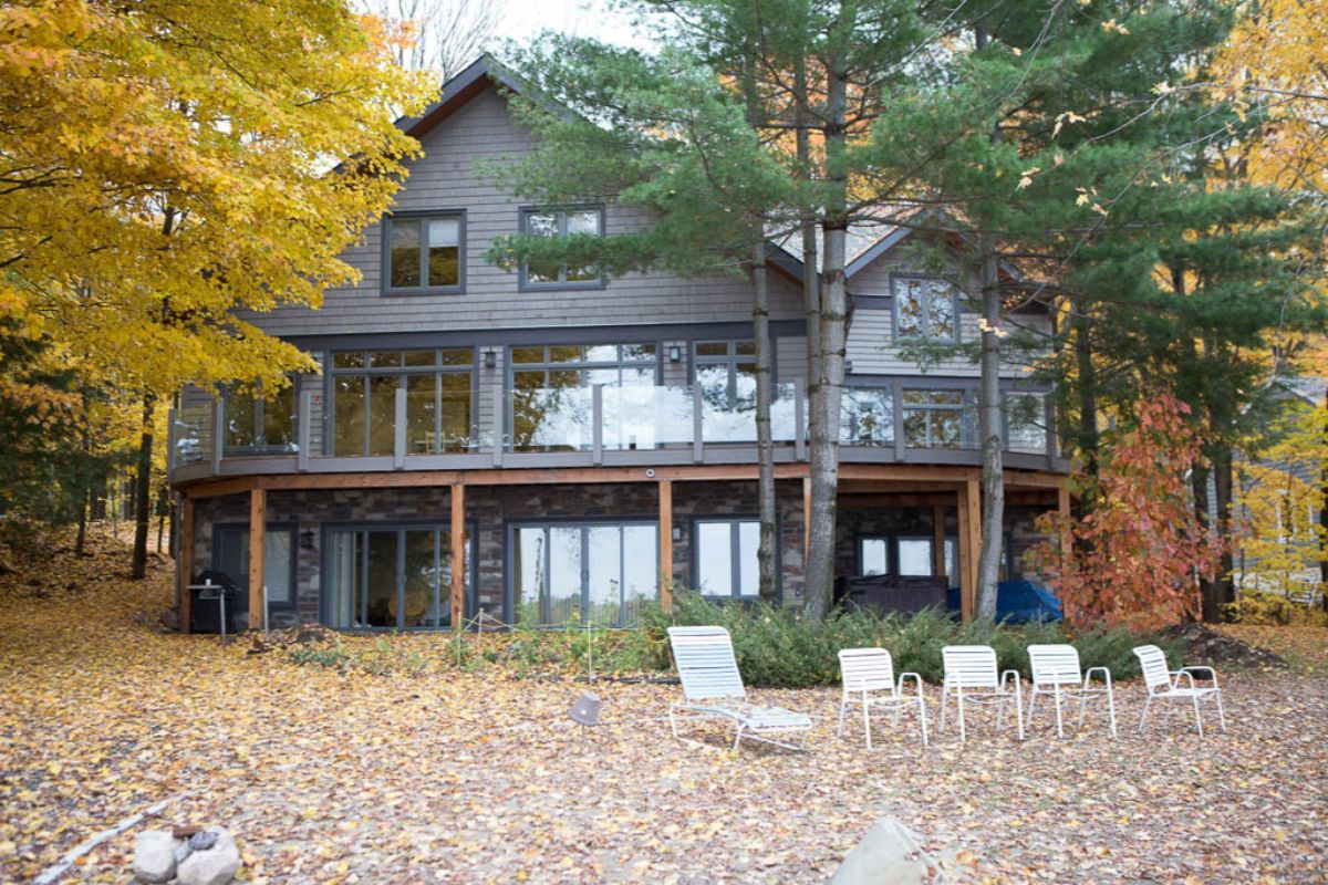 Custom Muskoka Cottage featuring a  large house with a lot of windows is surrounded by trees and leaves.