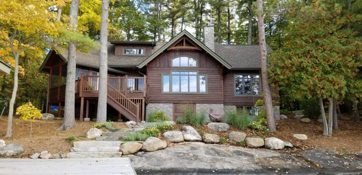 Custom Muskoka Cottage featuring a  large log cabin is sitting on the shore of a lake surrounded by trees.