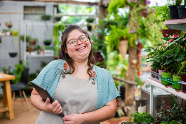 A woman with down syndrome is standing in a greenhouse holding a tablet and smiling.
