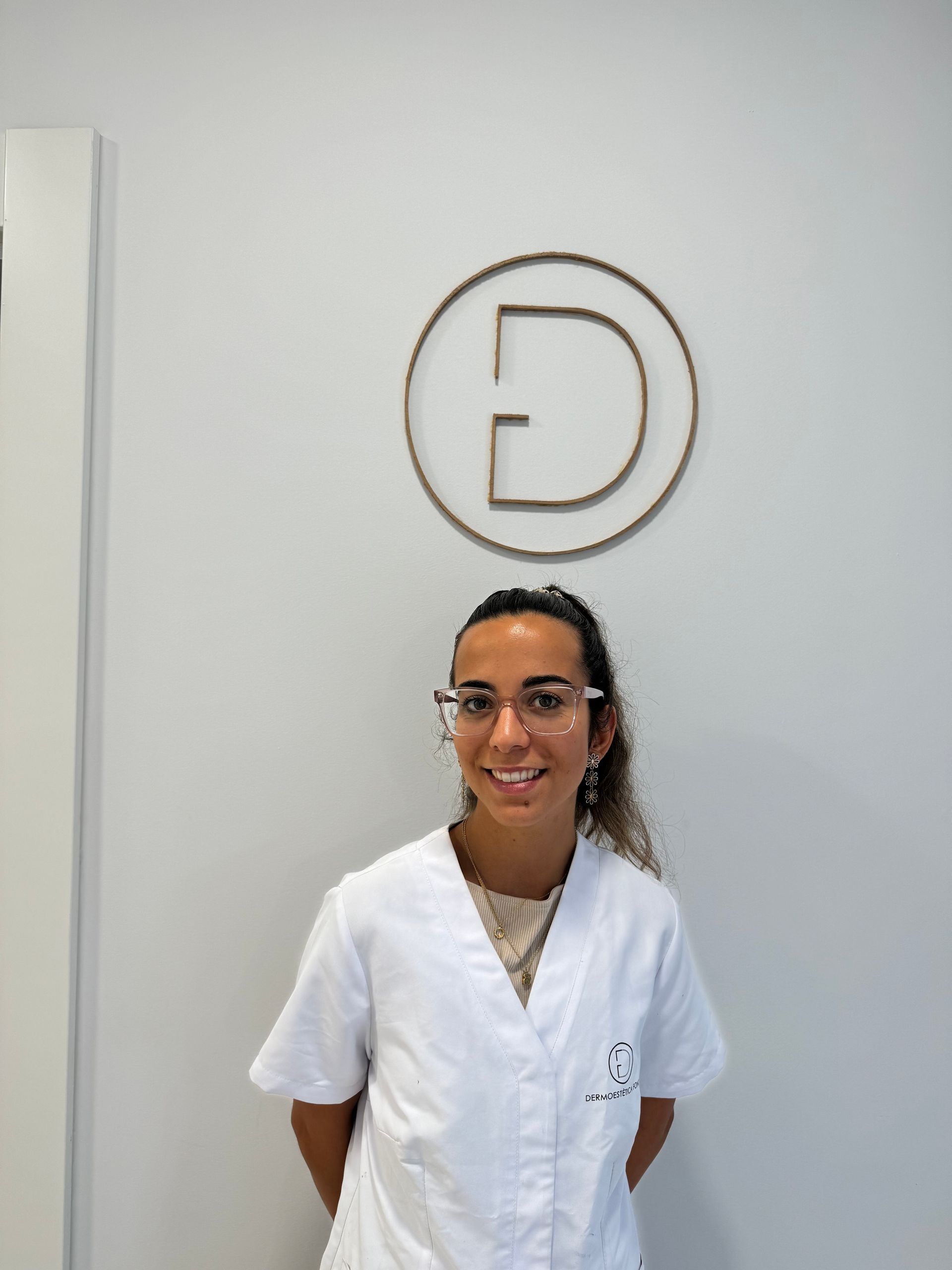 Una mujer con una bata blanca y gafas está parada frente a una pared con una letra d en ella.