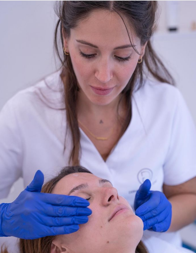 Mujer con guantes azules realizando un tratamiento facial a otra persona.