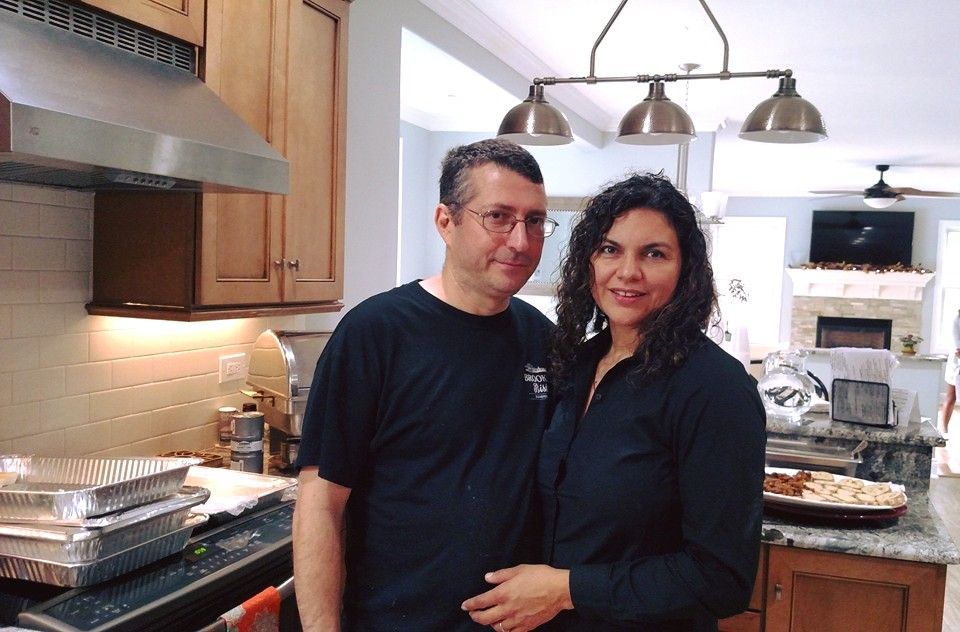 Couple in a kitchen, posing together. Kitchen has a stove, oven, and food containers.
