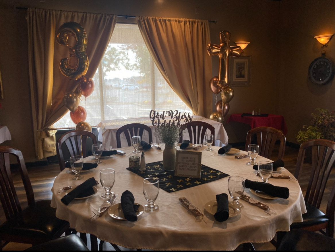 Round table set for a celebration with gold and orange balloons, a gold cross balloon, and window with curtains.