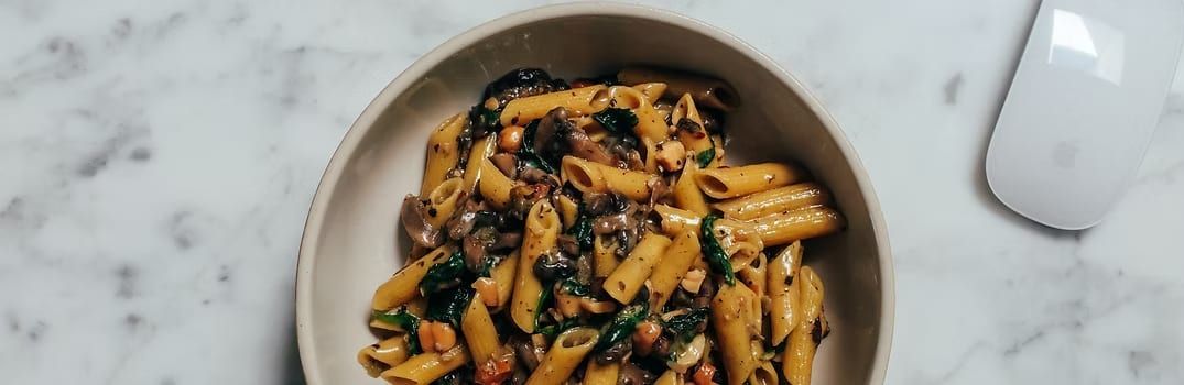 Penne pasta dish with mushrooms and spinach in a bowl on a marble surface, next to a computer mouse.