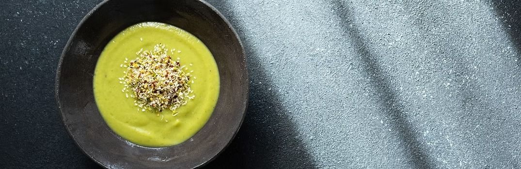 Green soup topped with seeds in a dark bowl on a textured surface.