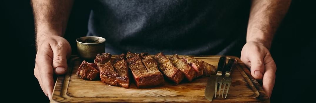 Man holding a wooden board with sliced steak and a small cup. A fork is on the board.