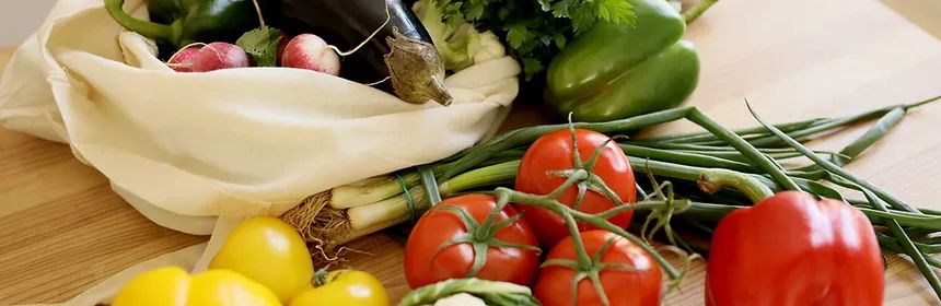 Fresh vegetables spilling from a reusable cloth bag onto a wooden surface: tomatoes, peppers, green onions.