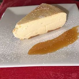 Slice of cheesecake with caramel sauce on a white plate, sprinkled with powdered sugar; red background.