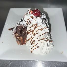 Dessert slice topped with whipped cream, chocolate drizzle, and a cherry, served on a white square plate.