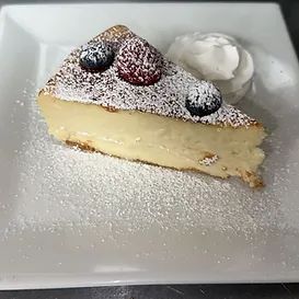 Slice of cheesecake with berries, powdered sugar, and whipped cream on a white plate.