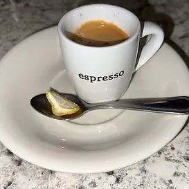 Espresso in white cup on saucer with spoon holding a lemon wedge.