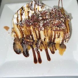 Dessert on a white plate: cheesecake with caramel, chocolate sauce, pecans, and whipped cream.