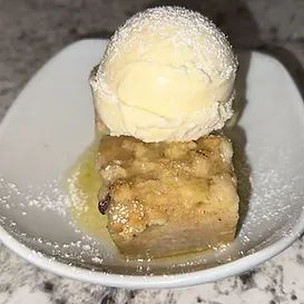 Bread pudding with vanilla ice cream in a white bowl, drizzled with sauce and dusted with powder.