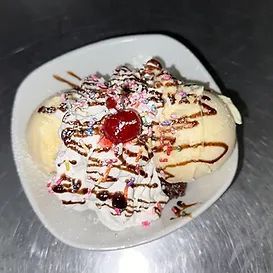 Banana split in white bowl with ice cream, whipped cream, sprinkles, cherry, and chocolate drizzle.