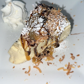 Slice of cheesecake, topped with caramel, chocolate, coconut flakes, and powdered sugar, with whipped cream.