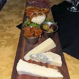 Wooden board with assorted cheeses, olives, sun-dried tomatoes, and bread, on a gold tablecloth.