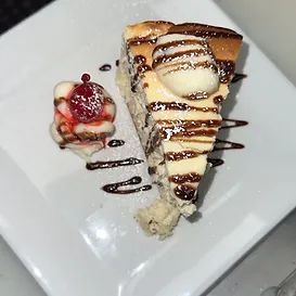 Slice of cheesecake drizzled with chocolate and topped with ice cream, served with a side of cream and cherry.