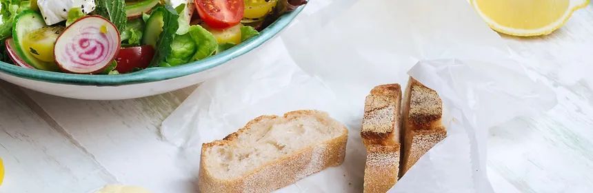 A fresh salad with bread and a lemon wedge on a white surface. The salad is in a teal-rimmed bowl.