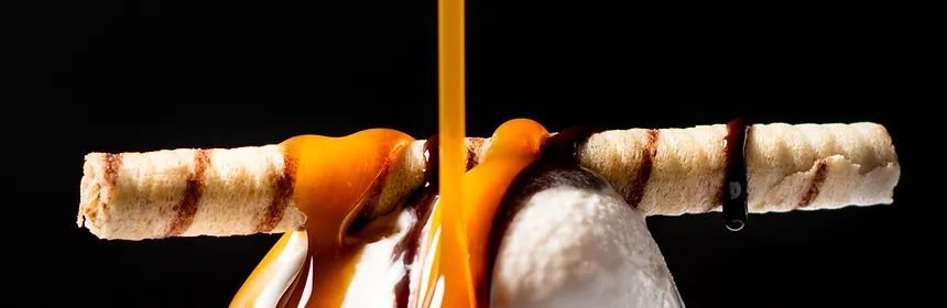 Caramel being poured over ice cream, wafer and chocolate against a black background.