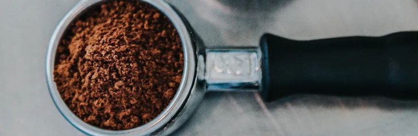 Close-up of a portafilter filled with coffee grounds against a silver metal surface; a black handle is attached.