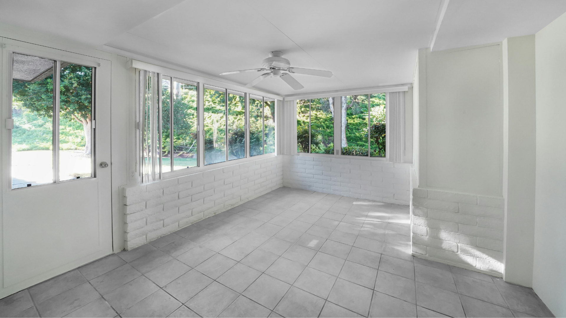 Bright, empty sunroom with white walls, tiled floor, and many windows overlooking greenery. A ceiling fan is visible.