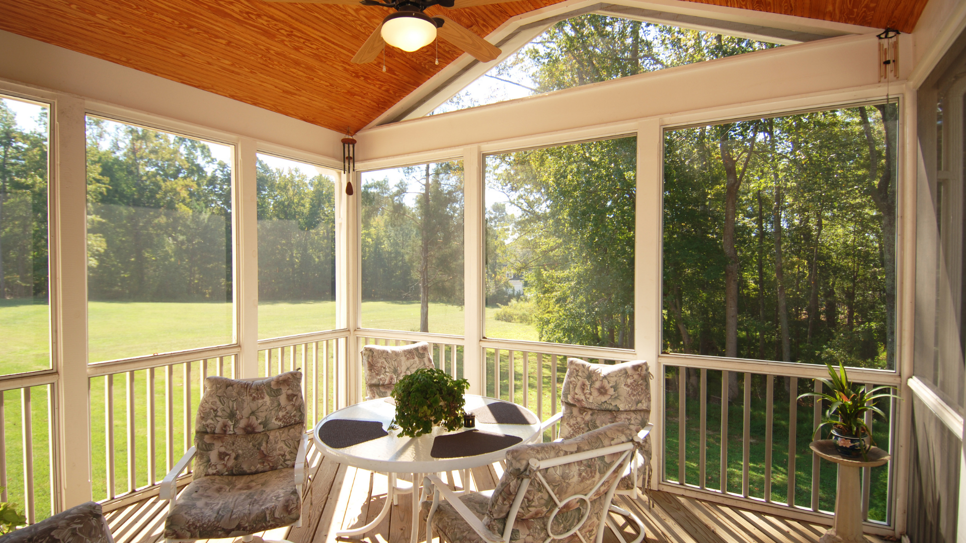 Screened porch with white furniture, looking out onto a green lawn and trees, with a wood ceiling.