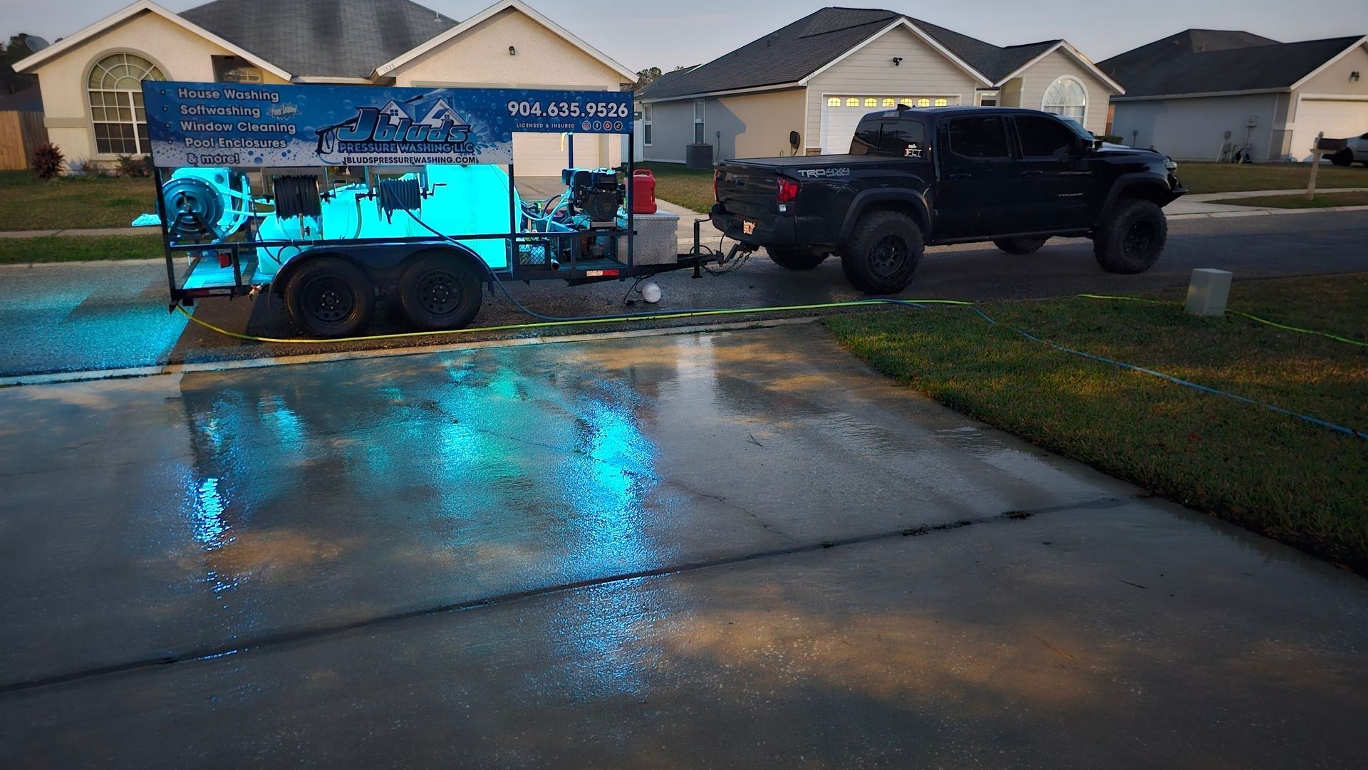 Black truck towing a trailer with blue glowing lights, parked on a driveway in front of suburban houses.