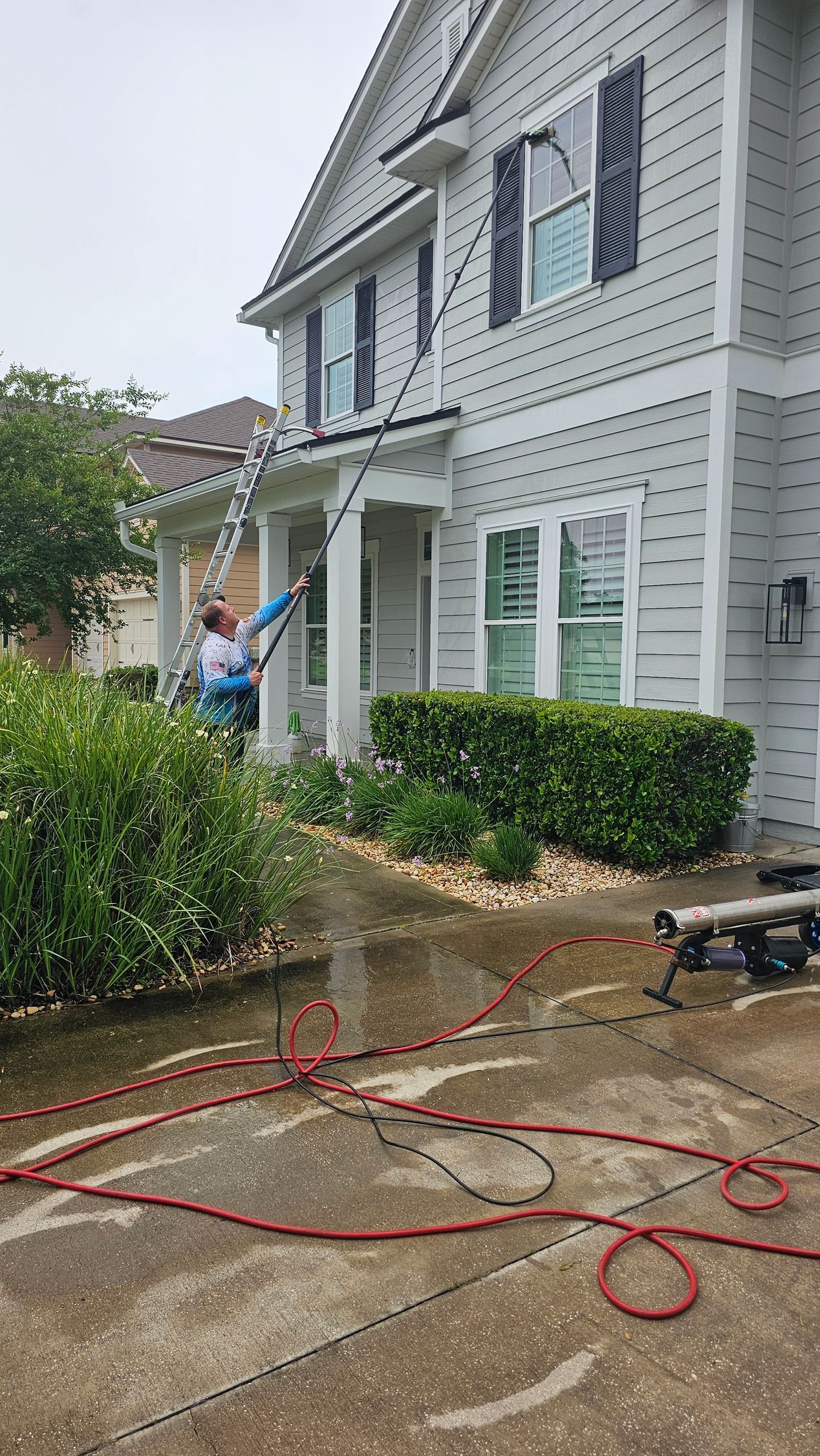 Person power washing a two-story house. Red hoses on concrete. Green bushes in front.