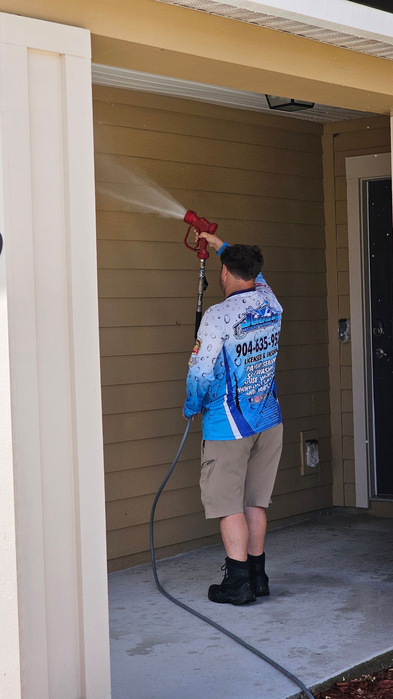 A man in blue shirt power washes the side of a beige building. Gray concrete floor.