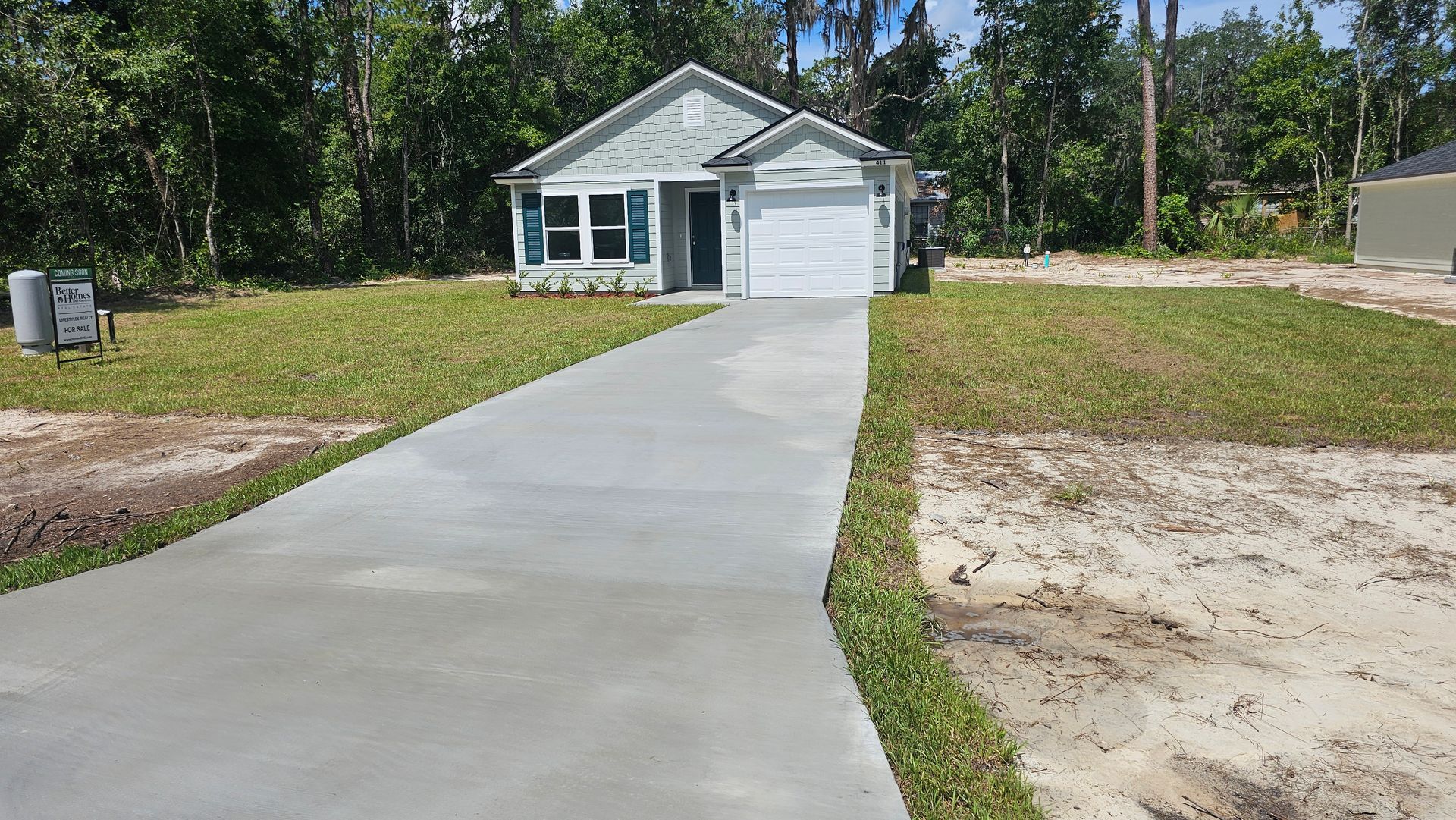 Small house with white garage and blue shutters, concrete driveway, grassy yard, and trees in the background.