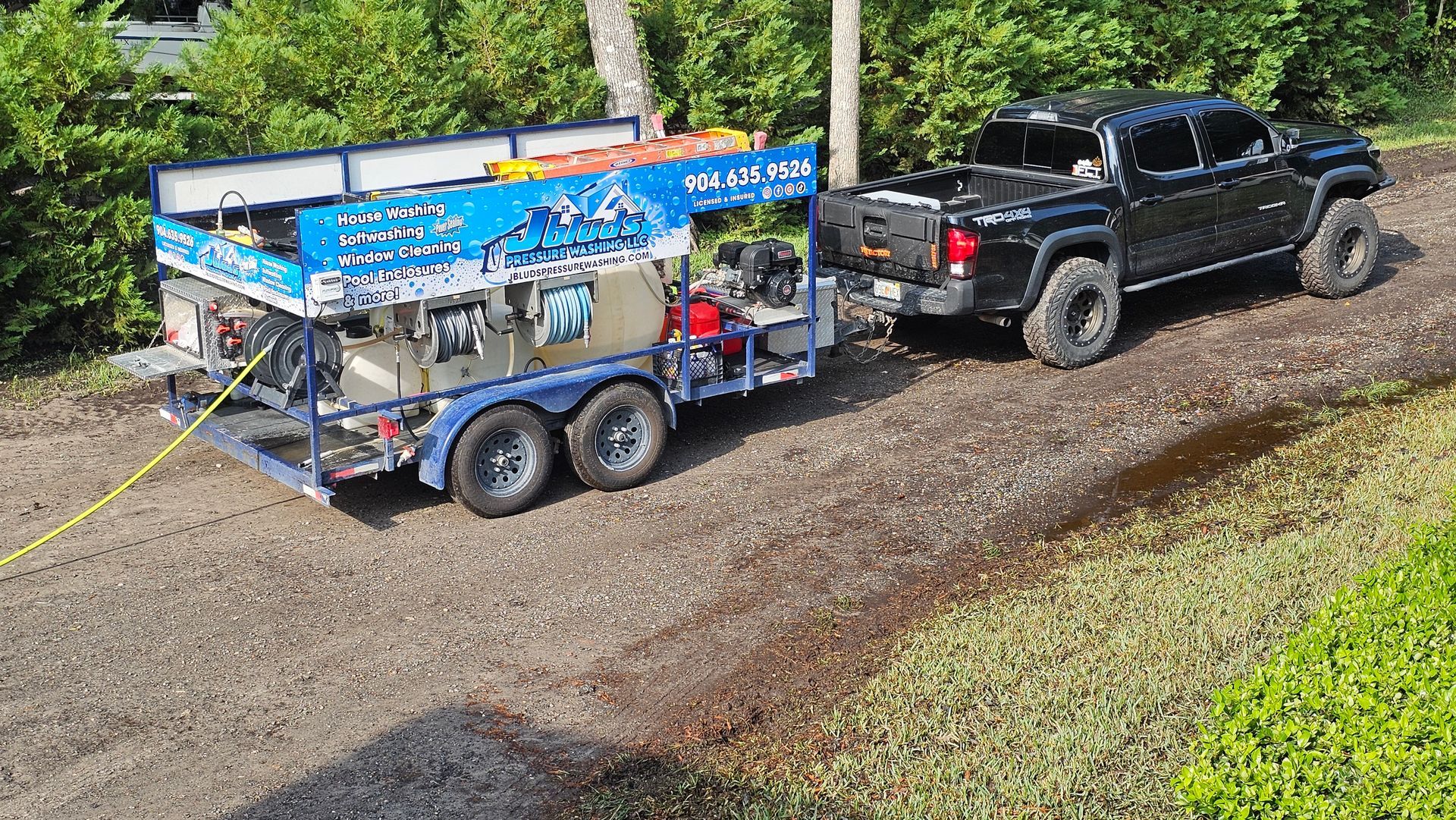 Black truck towing a trailer with pressure washing equipment on a gravel driveway near greenery.
