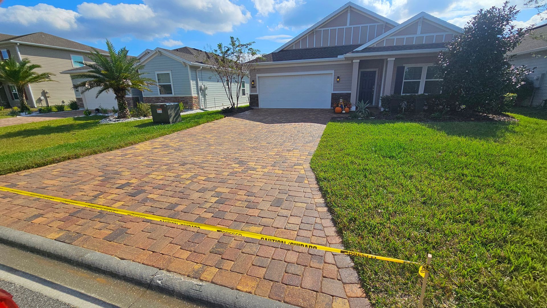 Brick driveway leading to a beige house with a two-car garage, bordered by green grass on a sunny day.