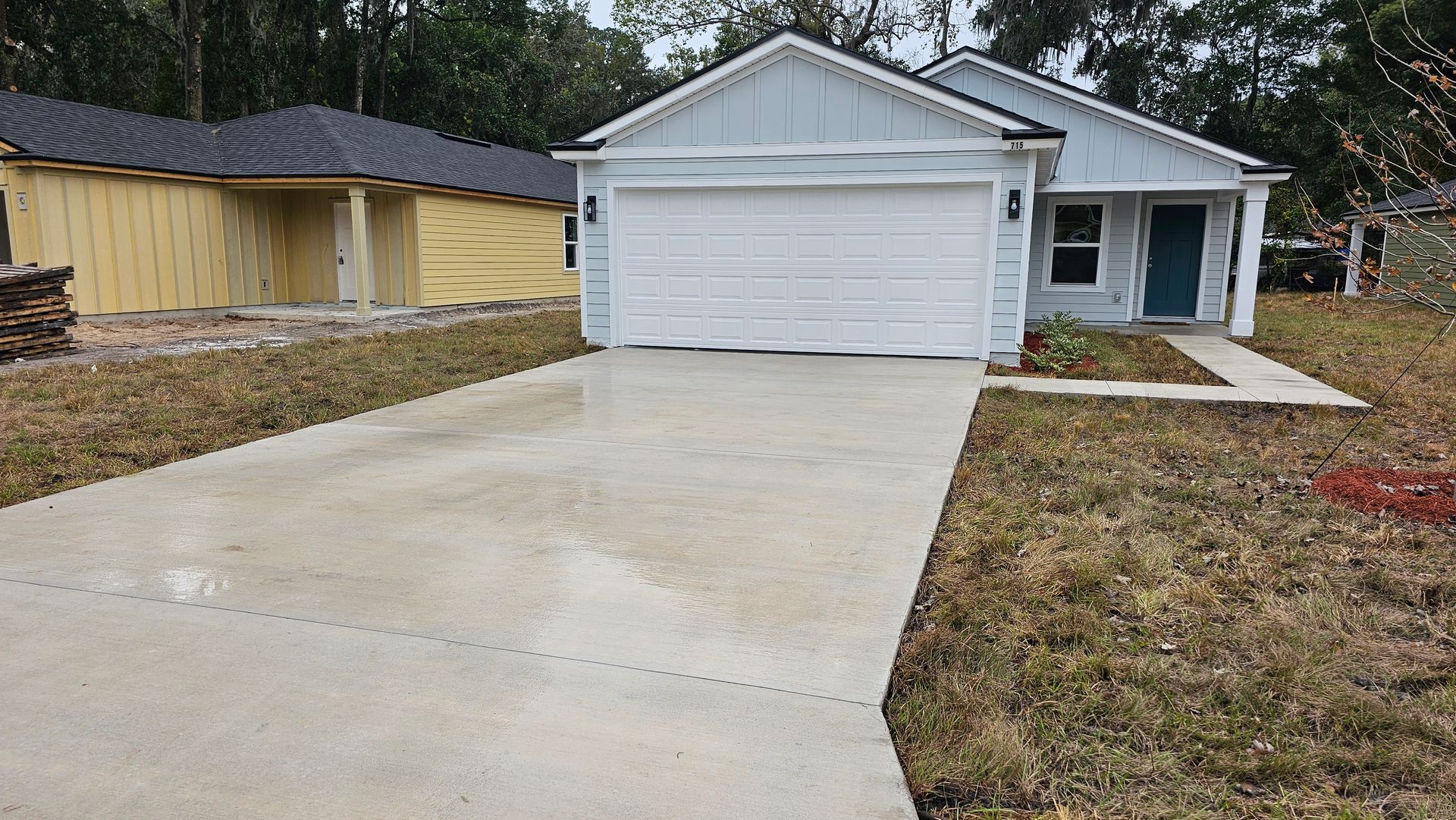 A light blue house with a white garage door and a concrete driveway. Yellow house in background.