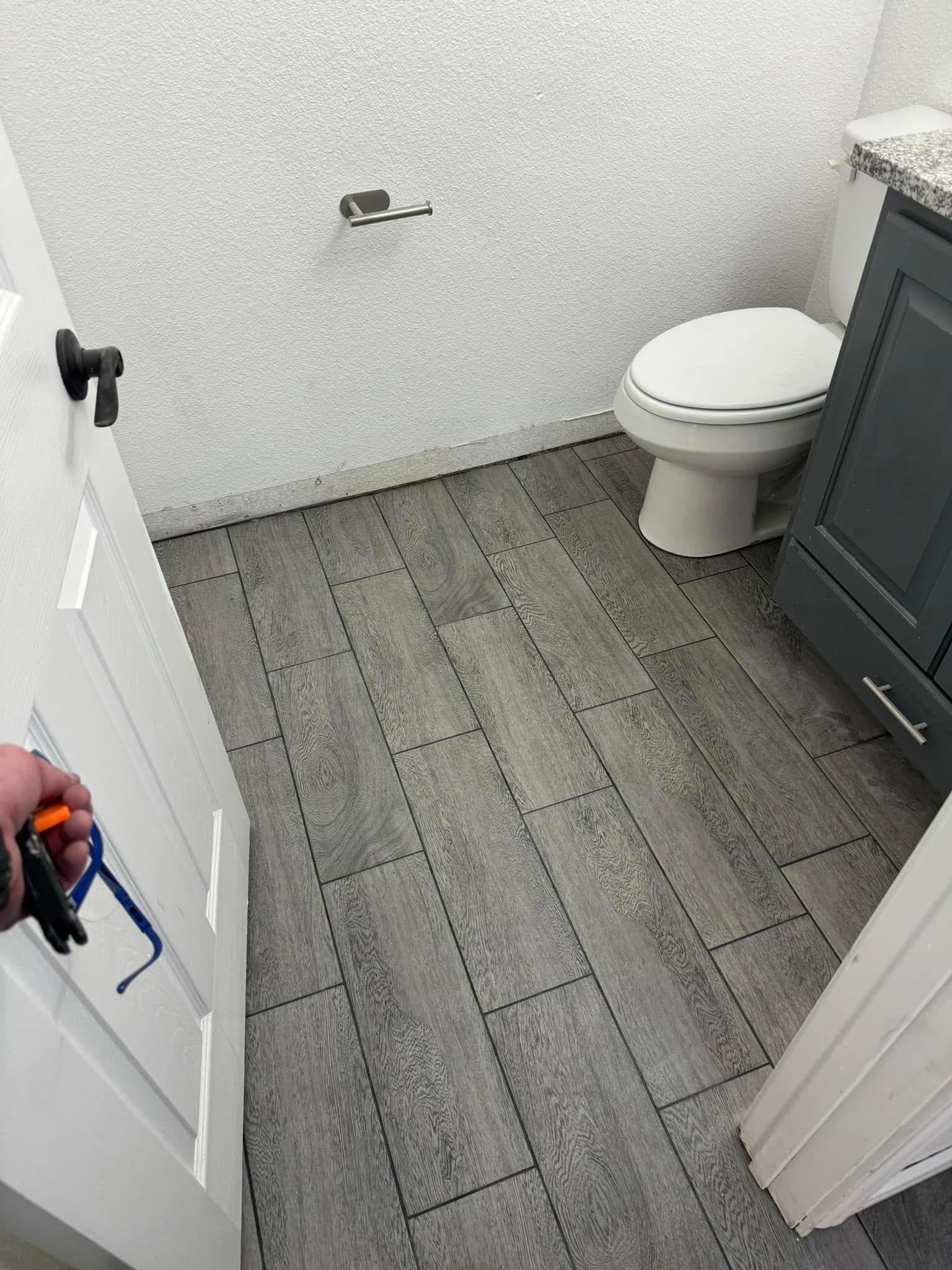 Bathroom interior: Gray tile floor, toilet, vanity, textured white wall, partially open white door with hardware.