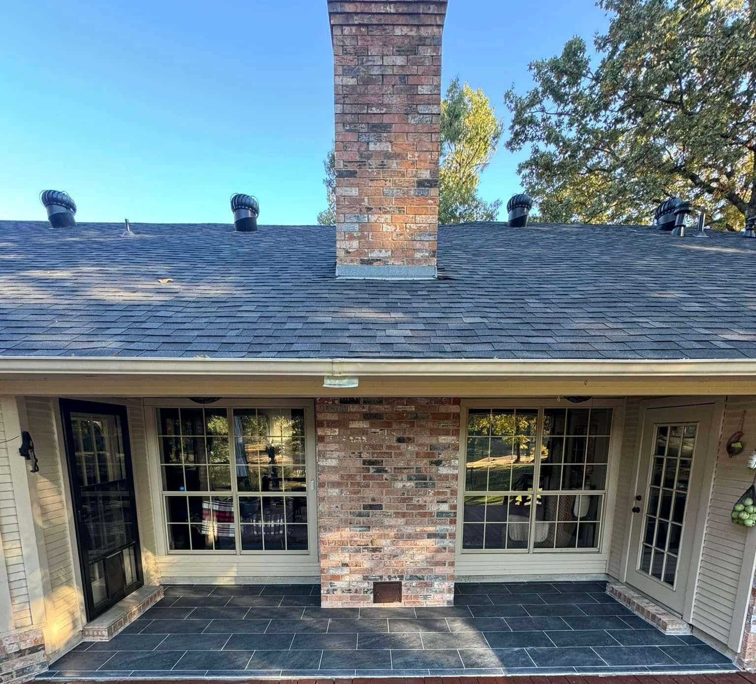Exterior view of a brick chimney on a dark shingled roof, windows below, and a black brick patio.