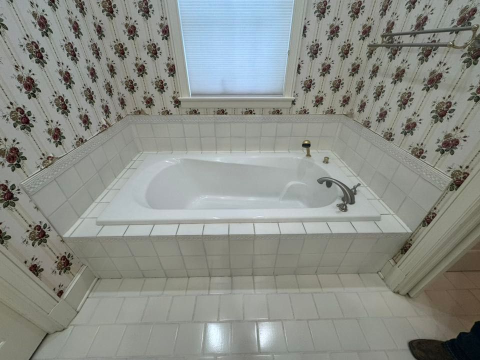 Built-in white bathtub in a bathroom, surrounded by white tile and floral wallpaper, with a window above.