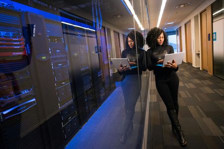 Femme en tenue noire, tenant un ordinateur portable, debout près des racks de serveurs dans un centre de données.