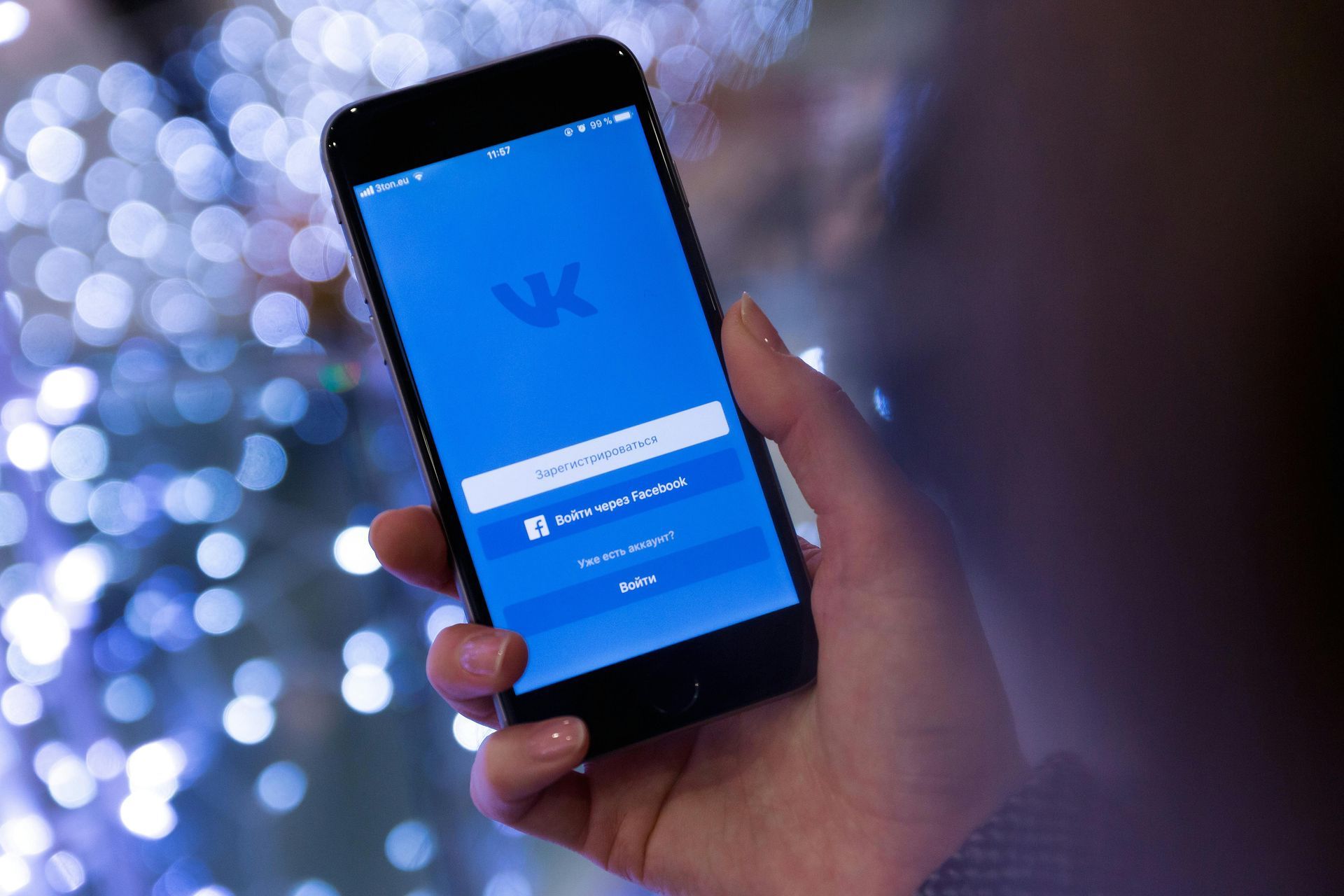Personne tenant un téléphone affichant l'écran de connexion VK, sur un fond flou et éclairé en bleu.