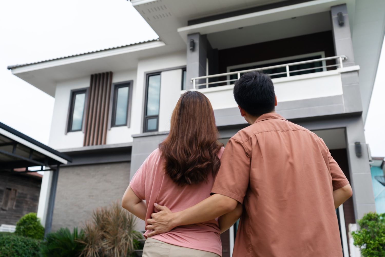 A couple stands with their arms around each other, looking up at their modern two-story house.