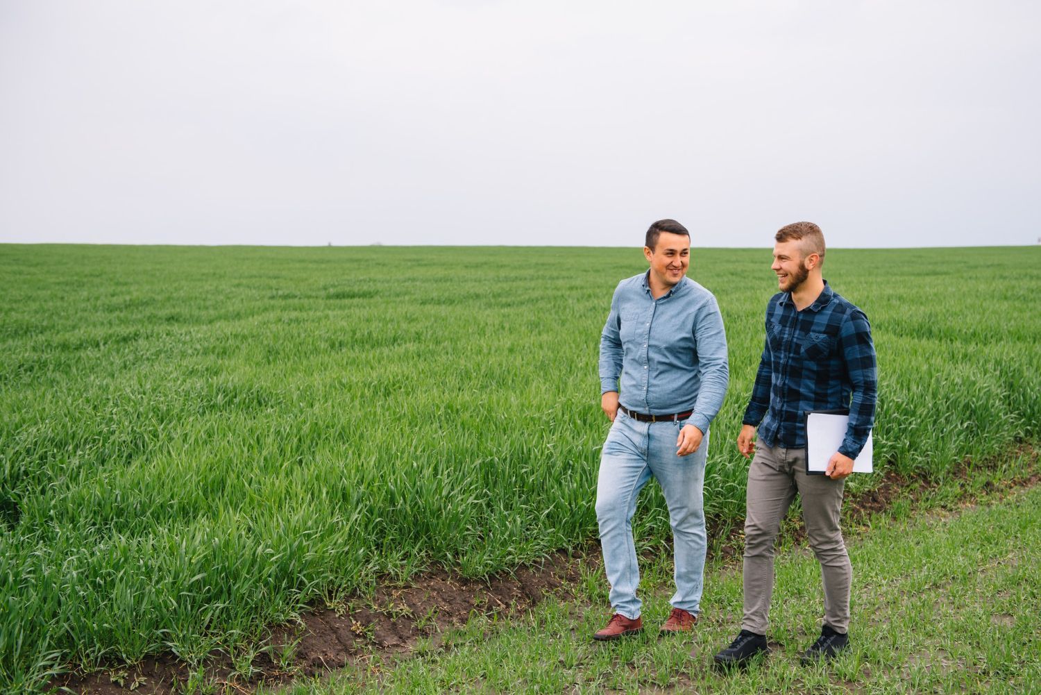 Two people walk through a green field, smiling and conversing while holding a clipboard.