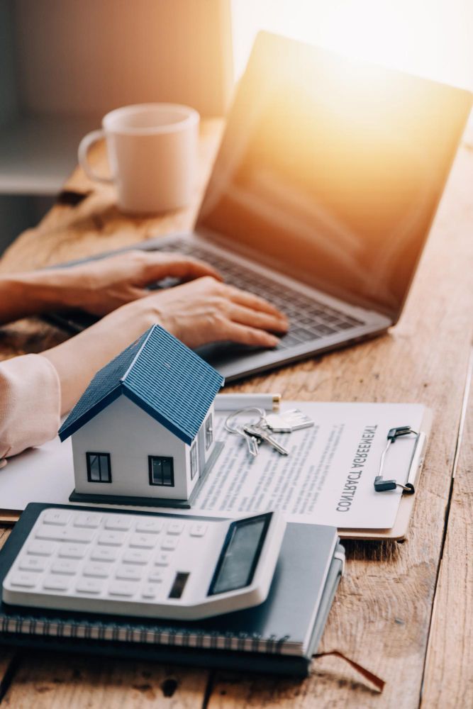 Hands typing on a laptop next to a house model, keys, a calculator, and documents on a wooden desk.