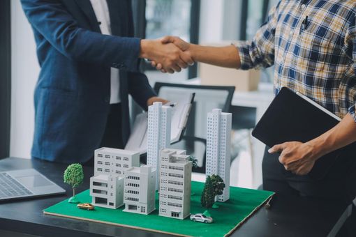 Two professionals shaking hands over a miniature architectural model of city buildings on a table.