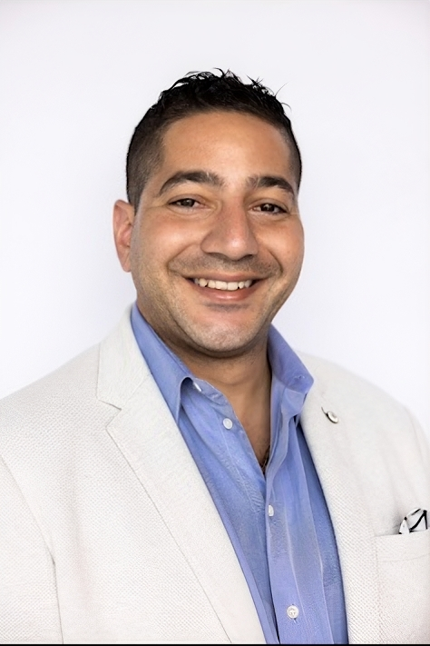A person smiling in a light-colored blazer over a blue collared shirt against a plain white background.