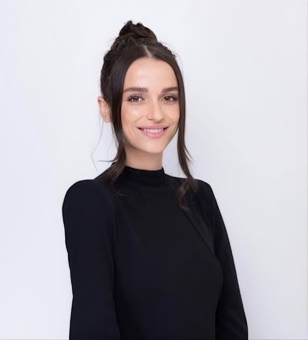 A person with dark hair pulled into a top knot smiling at the camera, wearing a black long-sleeved shirt against a white.
