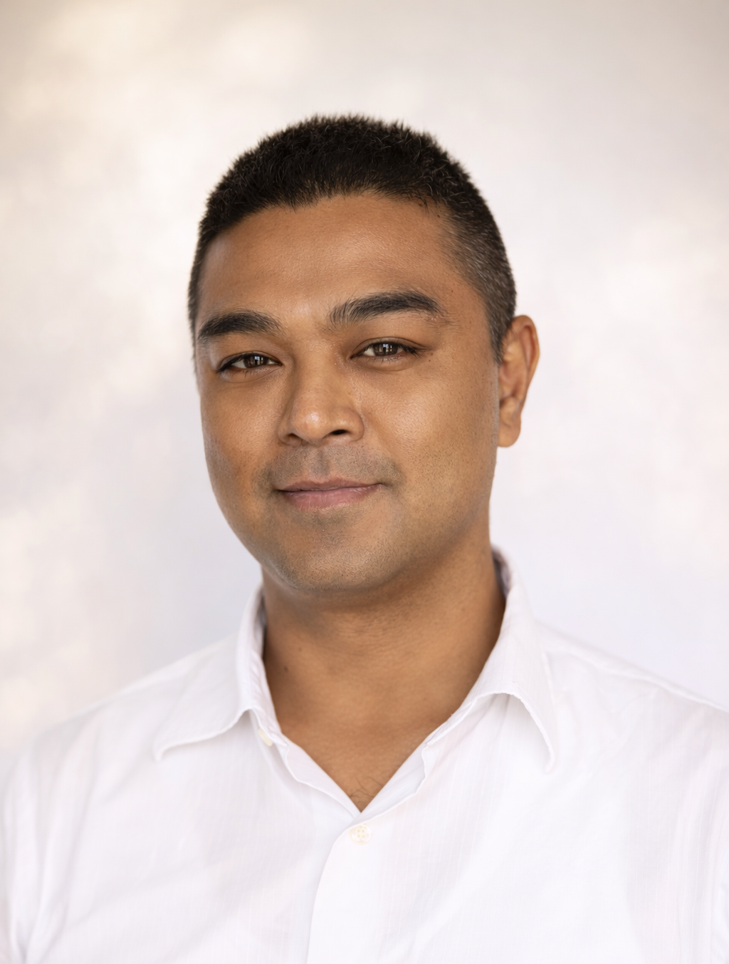 A person with short, dark hair and a neutral expression, wearing a white collared shirt against a soft, light background.