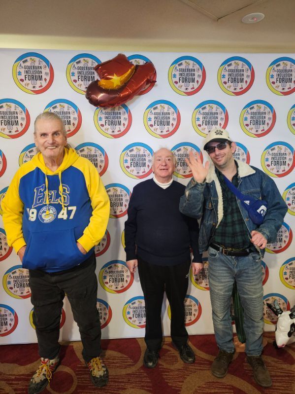 Three men pose in front of a colorful backdrop, one waving. Cowboy hat balloon floats above them.